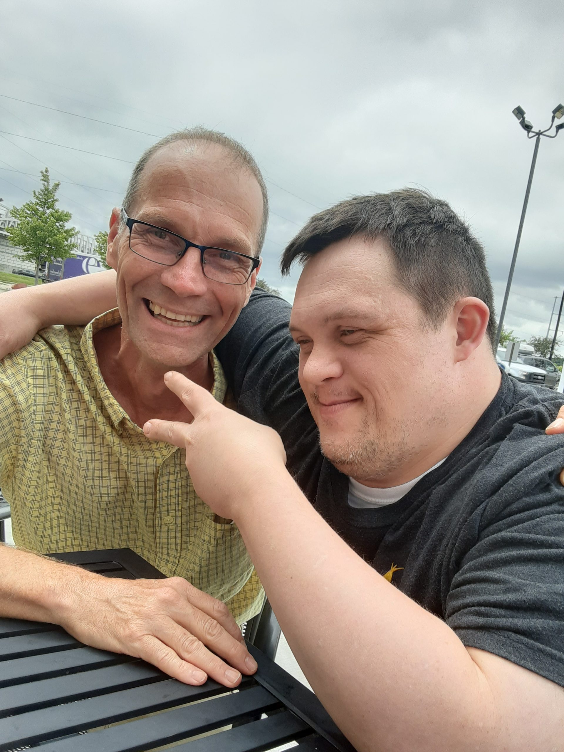 Two smiling men. The man on the left is looking at the camera and is wearing glasses and a yellow plaid shirt. The man on the right has Down syndrome and is wearing a gray t shirt.
